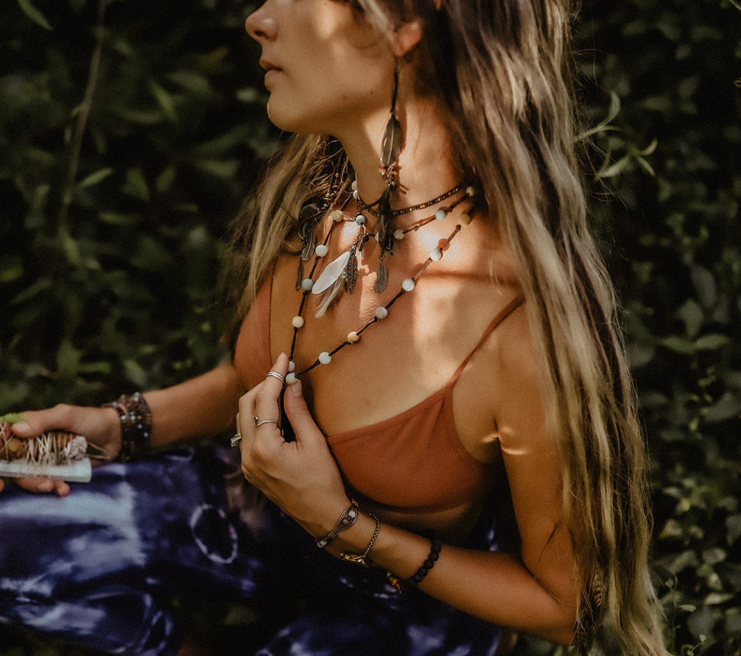A woman in a bohemian style sits among green foliage, wearing a rust-colored camisole, blue and white tie-dye pants, layered necklaces, and a patterned headband.